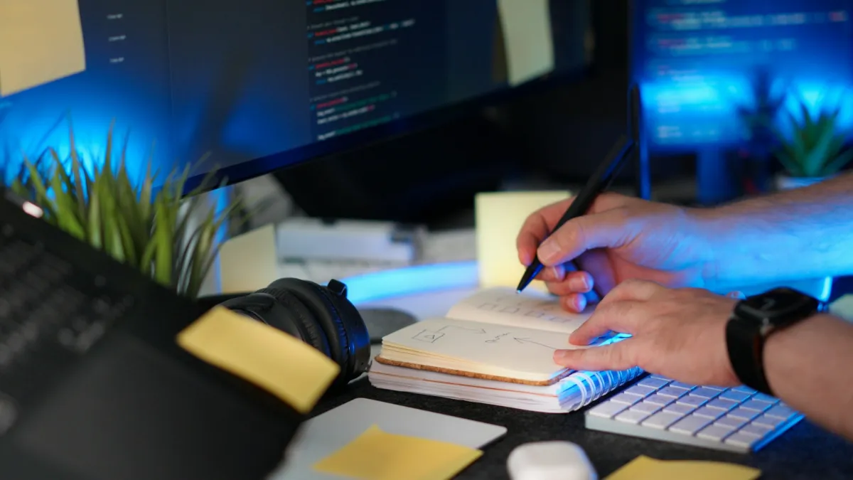 A developer writing a plan in a notebook beside monitors displaying code